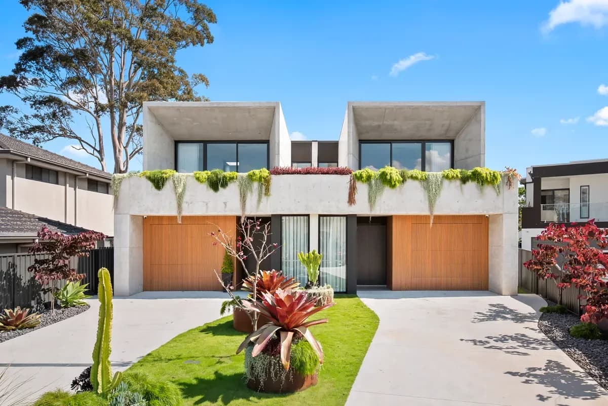 Finished daytime facade of the paired duplex with concrete upper forms, timber garage doors, and established planting