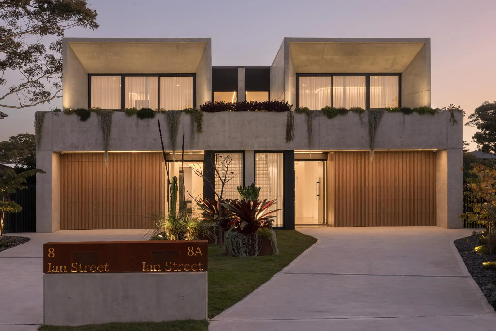 Street view of the Ian Duplex with concrete upper forms, timber garage doors, and landscaped front gardens at dusk
