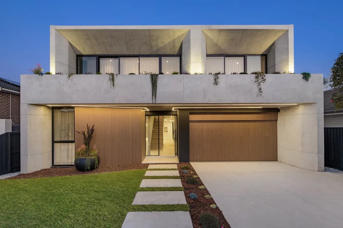 Sturdee Residence street elevation at dusk with a concrete upper level, timber garage doors, and a landscaped entry